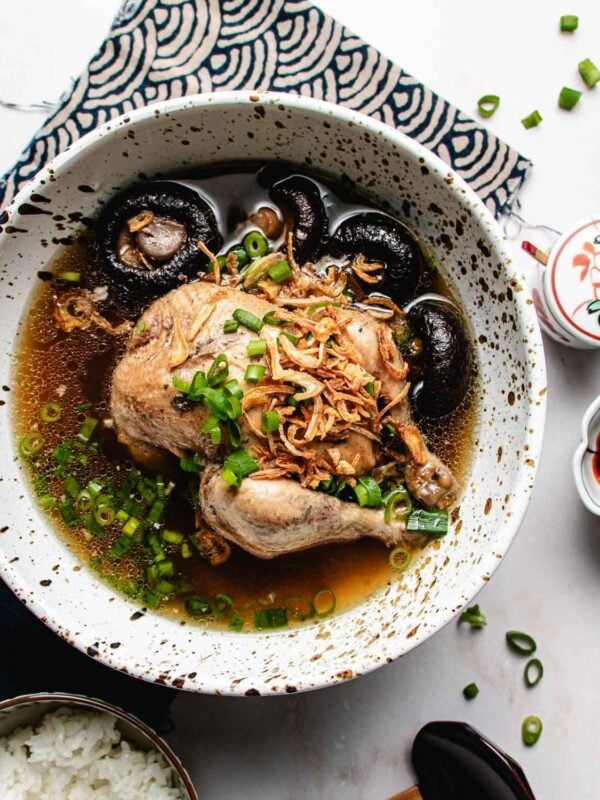 A bowl of Cornish Hen Soup with whole chicken, shiitake mushrooms, fried shallots, and chopped green onions in broth, served alongside white rice and dipping sauces on a patterned cloth.