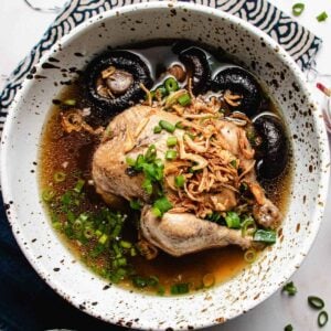 A bowl of Cornish Hen Soup with a whole chicken, shiitake mushrooms, chopped green onions, and crispy fried shallots in a dark, flavorful broth. The white bowl with black speckles sits on a patterned cloth.
