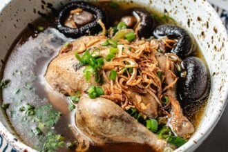 A bowl of Cornish Hen Soup features a whole chicken leg, shiitake mushrooms, chopped green onions, and crispy fried shallots in a clear broth.