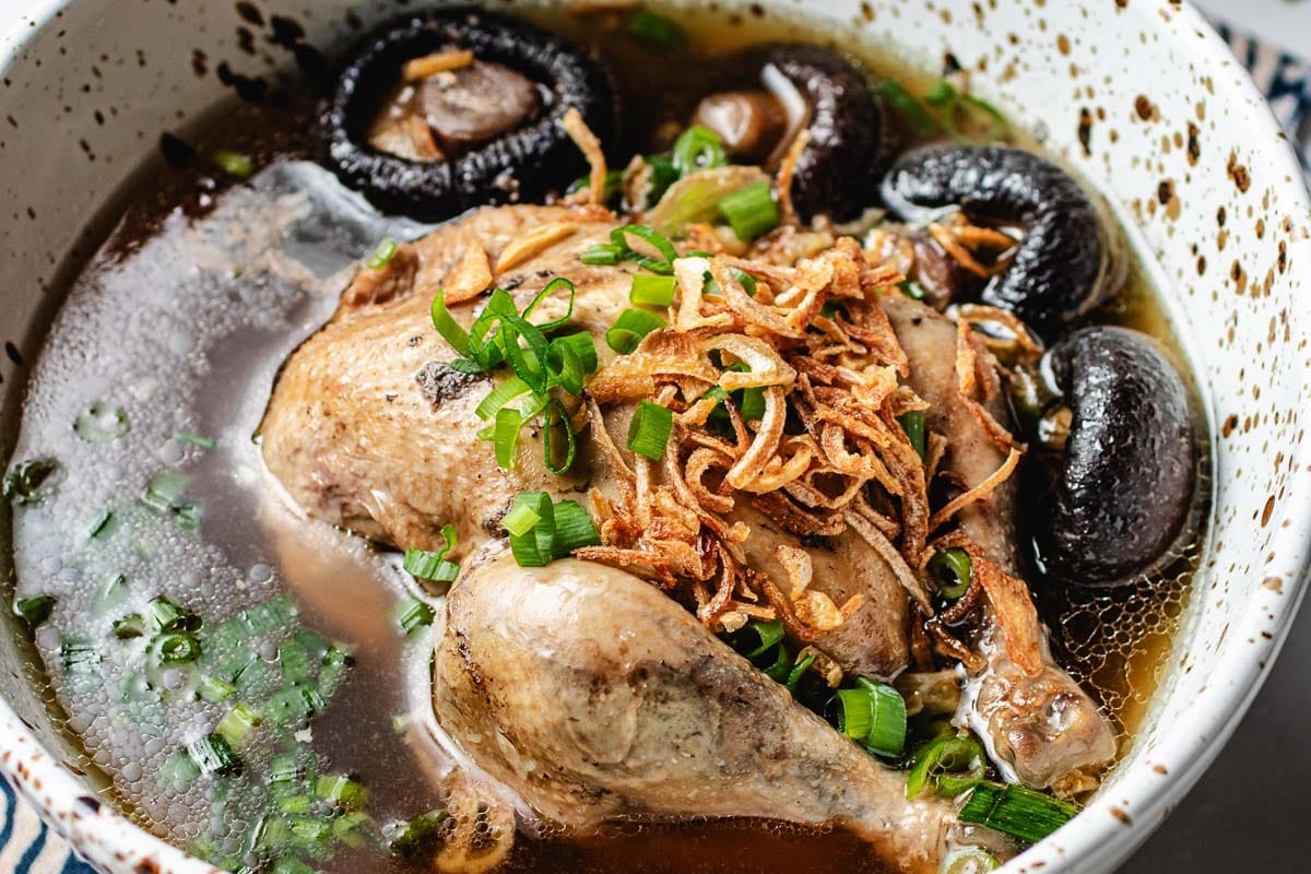 A bowl of Cornish Hen Soup features a whole chicken leg, shiitake mushrooms, chopped green onions, and crispy fried shallots in a clear broth.