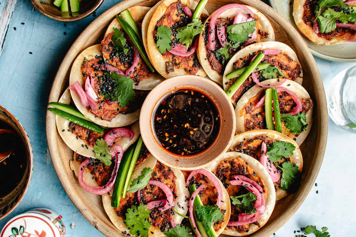 A plate of Smashed Dumpling Tacos—small dumpling wrappers topped with minced meat, pickled onions, cilantro, black sesame seeds, and sliced green chilies—arranged around a bowl of soy-based dipping sauce.