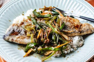 A plate with one piece of crispy pan seared rainbow trout served over white rice, topped with sautéed green onions and thinly sliced vegetables, with a fork and knife on the side.