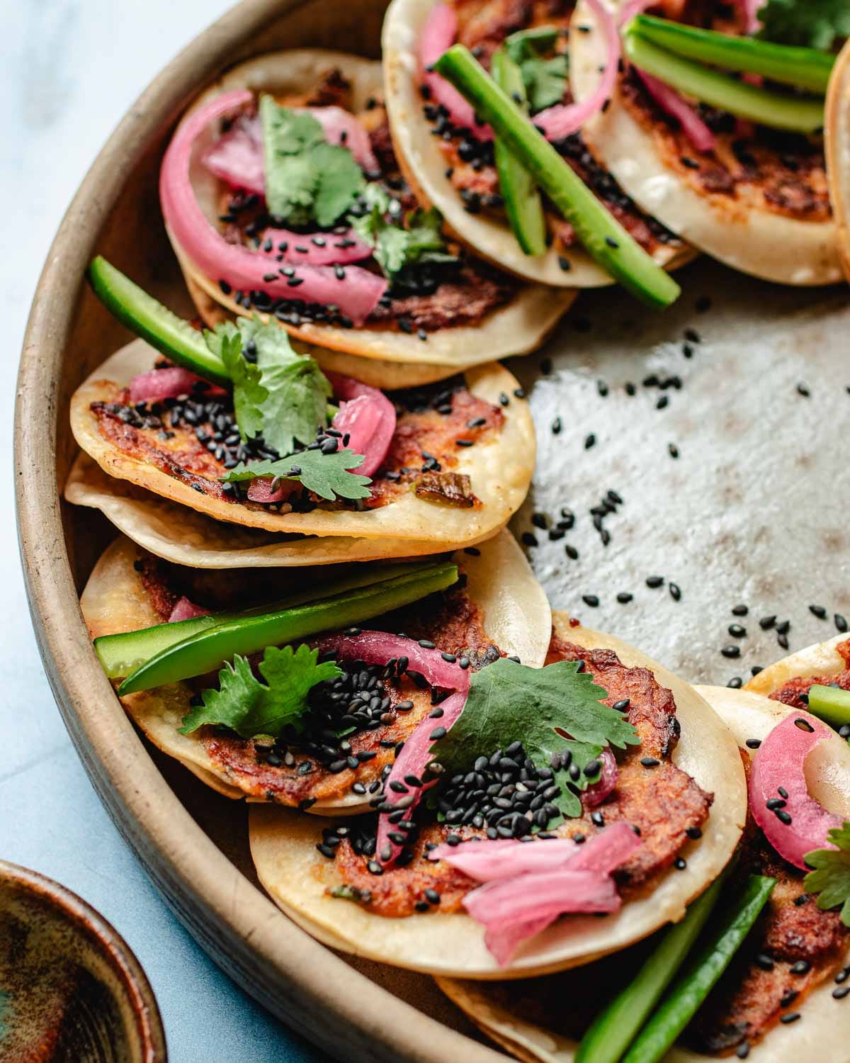 A plate of Smashed Dumpling Tacos, small and crispy, topped with wonton minced meat, black sesame seeds, fresh cilantro, pickled red onions, and cucumber slices, arranged in a circular pattern on a wooden platter.