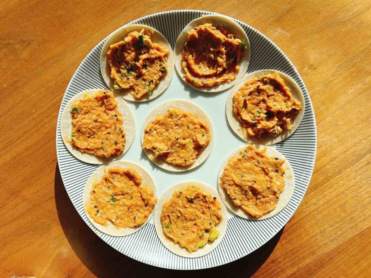 A white plate with black stripes around the rim holds eight small, round dumpling wrappers topped with chicken minced meat - Smashed Dumpling Tacos spread, arranged in a circle on a wooden table. Sunlight casts shadows across the scene.