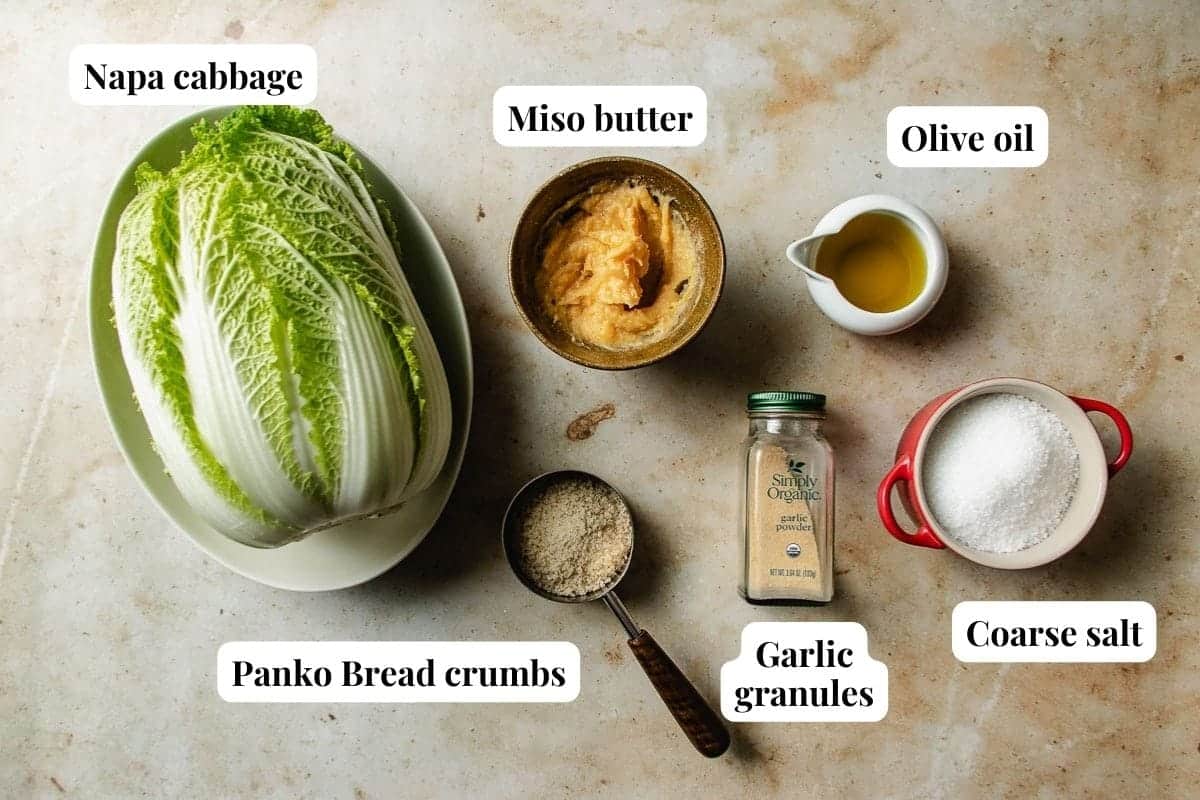 Top-down view of ingredients for roasted napa cabbage: napa cabbage, a bowl of miso butter, olive oil in a small pitcher, a cup of coarse salt, garlic granules in a jar, and panko bread crumbs on a light surface, each labeled.