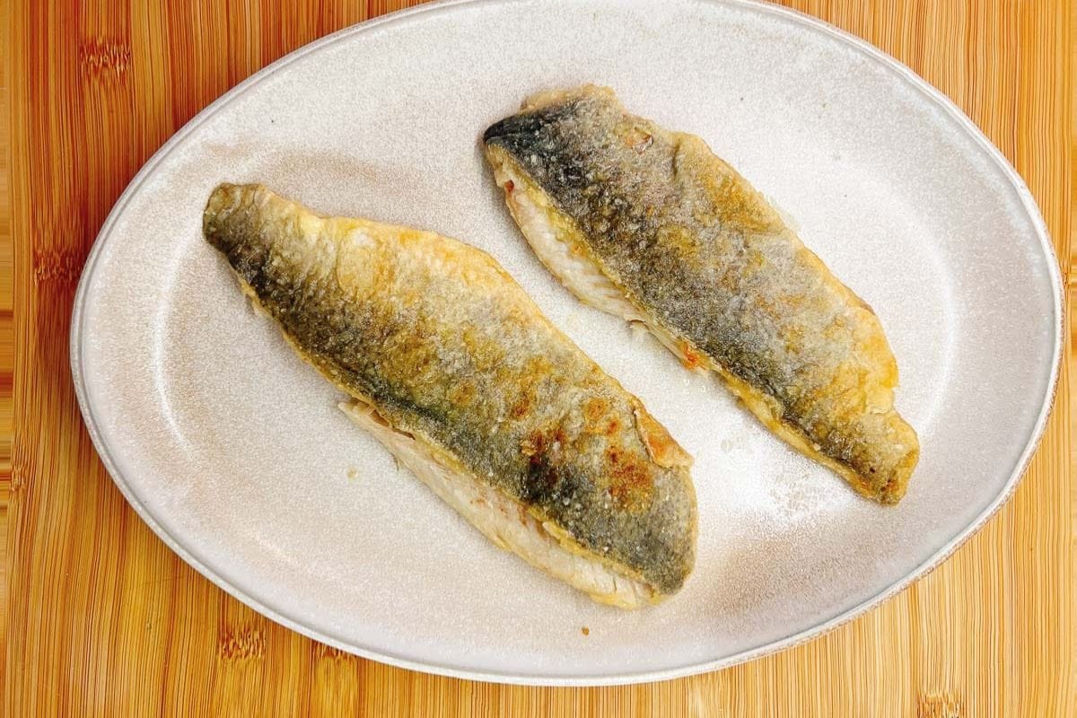 Two crispy pan fried trout fillets with golden skin are placed side by side on a white oval plate set on a wooden surface, showcasing an inviting pan fried trout presentation.