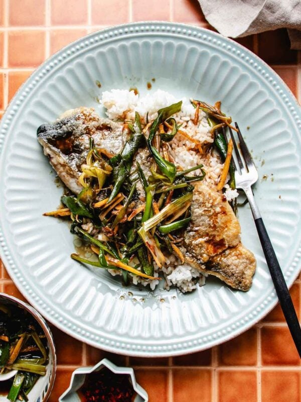 A plate of pan seared rainbow trout served over white rice, topped with sautéed green onions and thin strips of ginger, sits on a blue plate with a fork on an orange tiled surface. A small bowl of sauce is nearby.