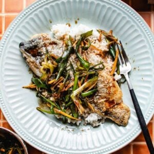 A plate with crispy pan fried trout fillets topped with sautéed green onions and ginger, served over white rice. A fork rests on the side of the light blue, round plate.