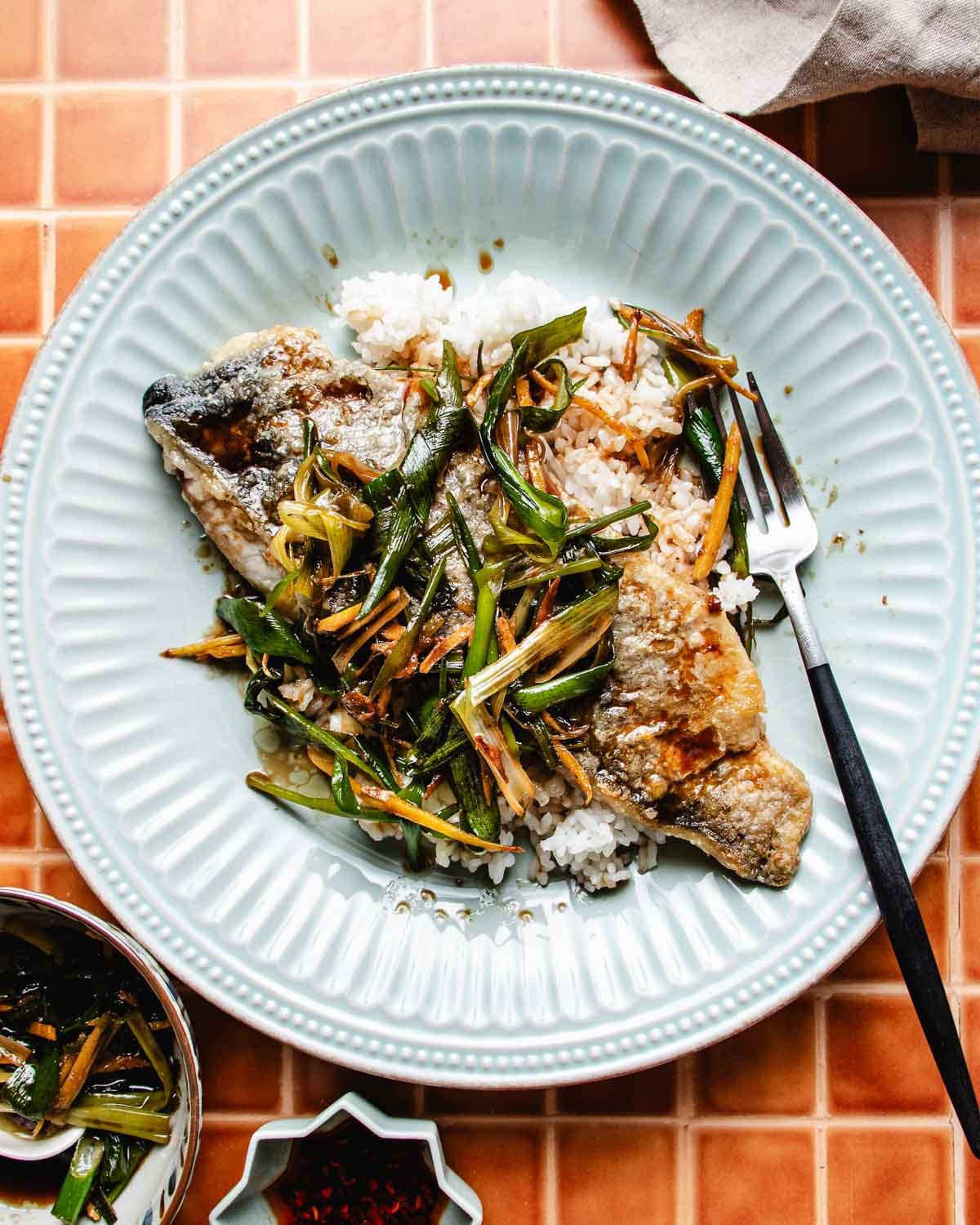 A plate of pan seared rainbow trout served over white rice, topped with sautéed green onions and thin strips of ginger, sits on a blue plate with a fork on an orange tiled surface. A small bowl of sauce is nearby.