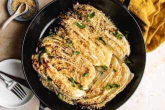 A skillet filled with roasted napa cabbage wedges topped with golden panko breadcrumbs and herbs. A spoon, small bowl of breadcrumbs, plates, and forks are nearby on the table.