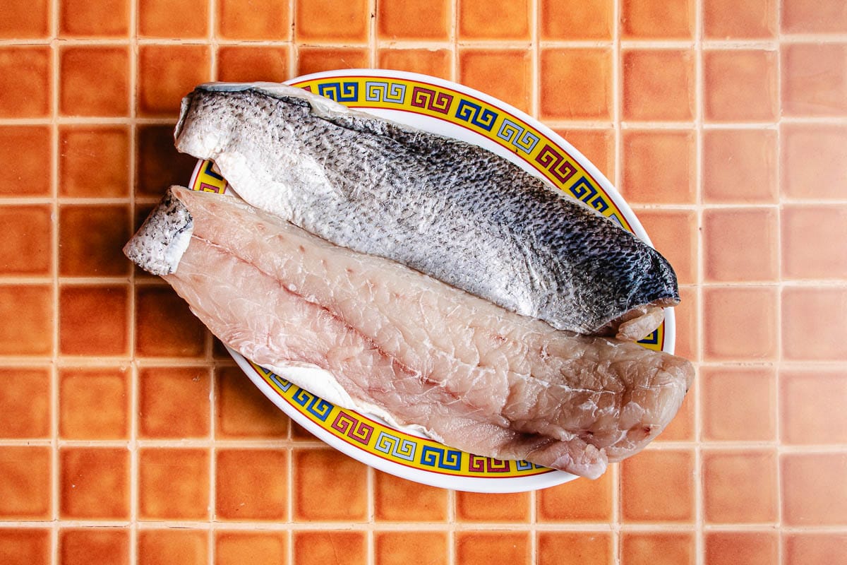 Two raw rainbow trout fillets, ideal for a pan fried trout or rainbow trout recipe, are placed side by side on an oval plate with a decorative border, set on an orange tiled surface.