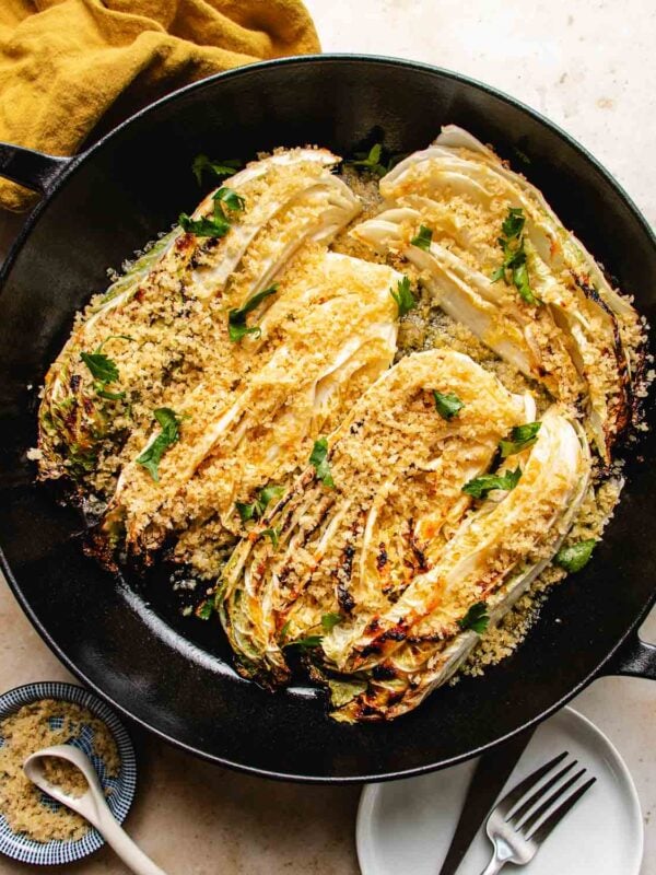 Roasted napa cabbage wedges, charred to perfection, are topped with golden panko breadcrumbs and fresh parsley in a black skillet, served with a side of extra breadcrumbs and a plate with utensils.