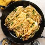 A black skillet filled with roasted napa cabbage wedges, topped with golden panko breadcrumbs and fresh herbs, sits on a light-colored surface with a small dish of extra breadcrumbs nearby.