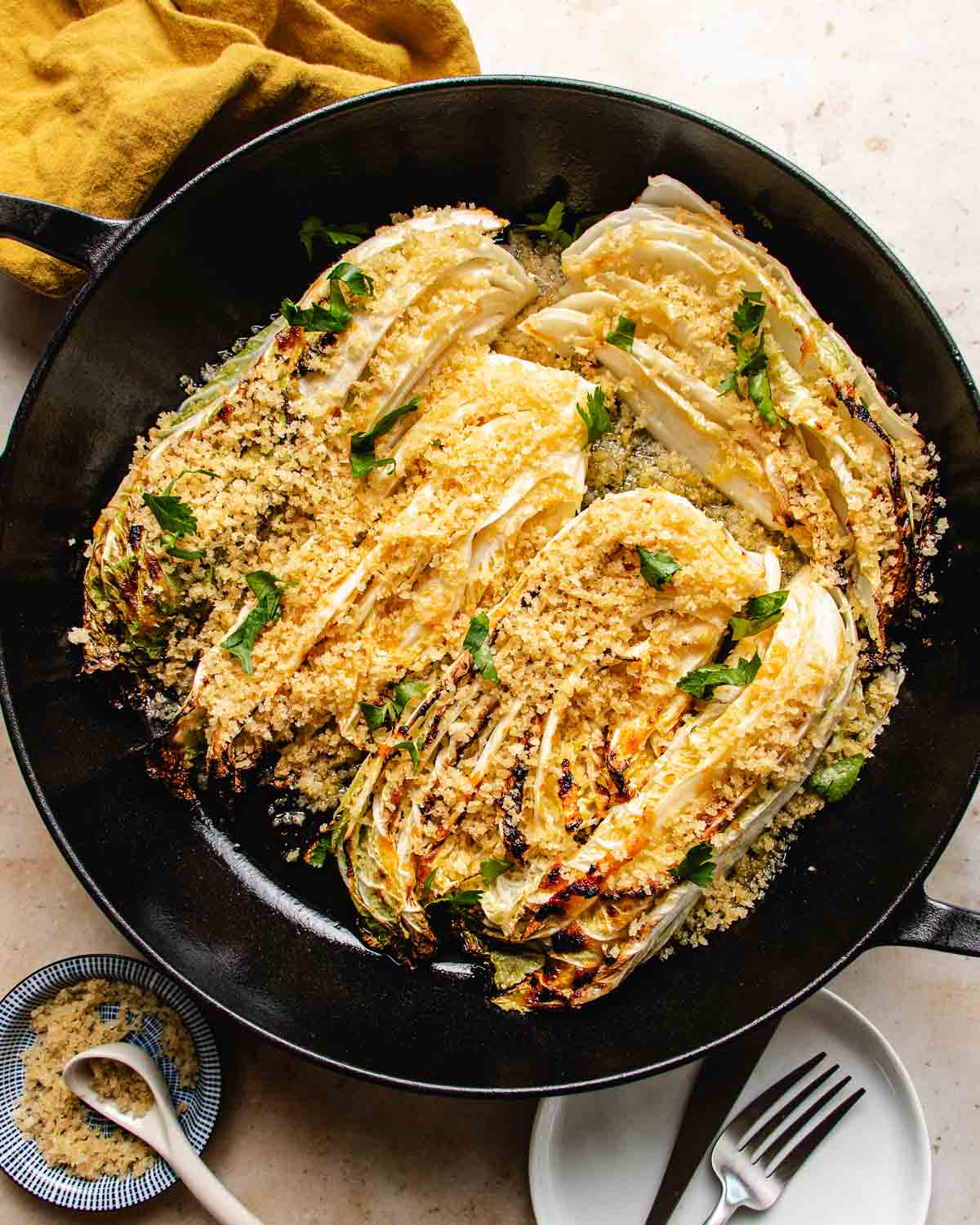 Roasted napa cabbage wedges, charred to perfection, are topped with golden panko breadcrumbs and fresh parsley in a black skillet, served with a side of extra breadcrumbs and a plate with utensils.