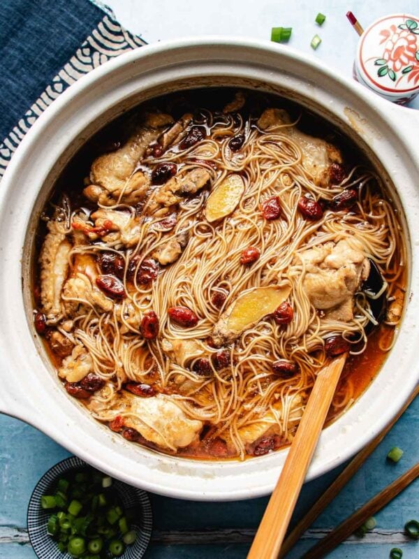 A large ceramic pot filled with simmering noodles, cut-up pieces of chicken, ginger slices, and goji berries in a rich brown sesame oil chicken soup broth, with a wooden ladle and scattered green onions nearby.
