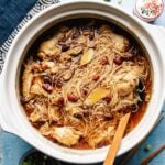 A ceramic clay pot filled with thin noodles, tender cut-up chicken pieces, and herbs in a rich sesame oil chicken soup broth, surrounded by chopsticks and a small plate on a blue surface.