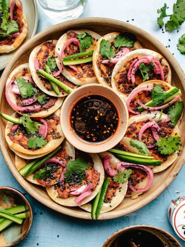 A round platter of mini dumpling wrappers topped with dumpling ground meat, cilantro, pickled onions, black sesame seeds, and cucumber slices surrounds a bowl of dark dipping sauce—perfect alongside Smashed Dumpling Tacos. Other dishes and utensils are partially visible on the table.