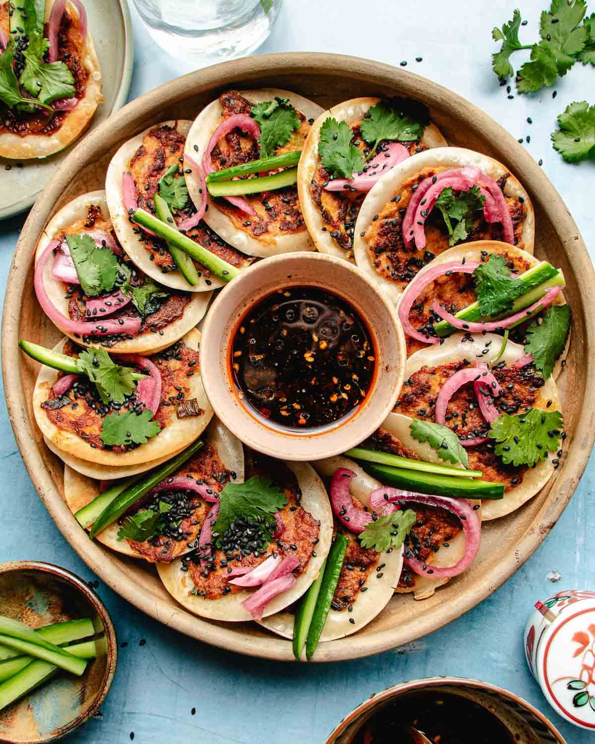 A round platter of mini dumpling wrappers topped with dumpling ground meat, cilantro, pickled onions, black sesame seeds, and cucumber slices surrounds a bowl of dark dipping sauce—perfect alongside Smashed Dumpling Tacos. Other dishes and utensils are partially visible on the table.