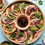 A wooden plate filled with small Smashed Dumpling Tacos topped with chicken wonton filling, cilantro, pickled onions, black sesame seeds, and sliced green peppers, all arranged around a bowl of dark dipping sauce in the center.
