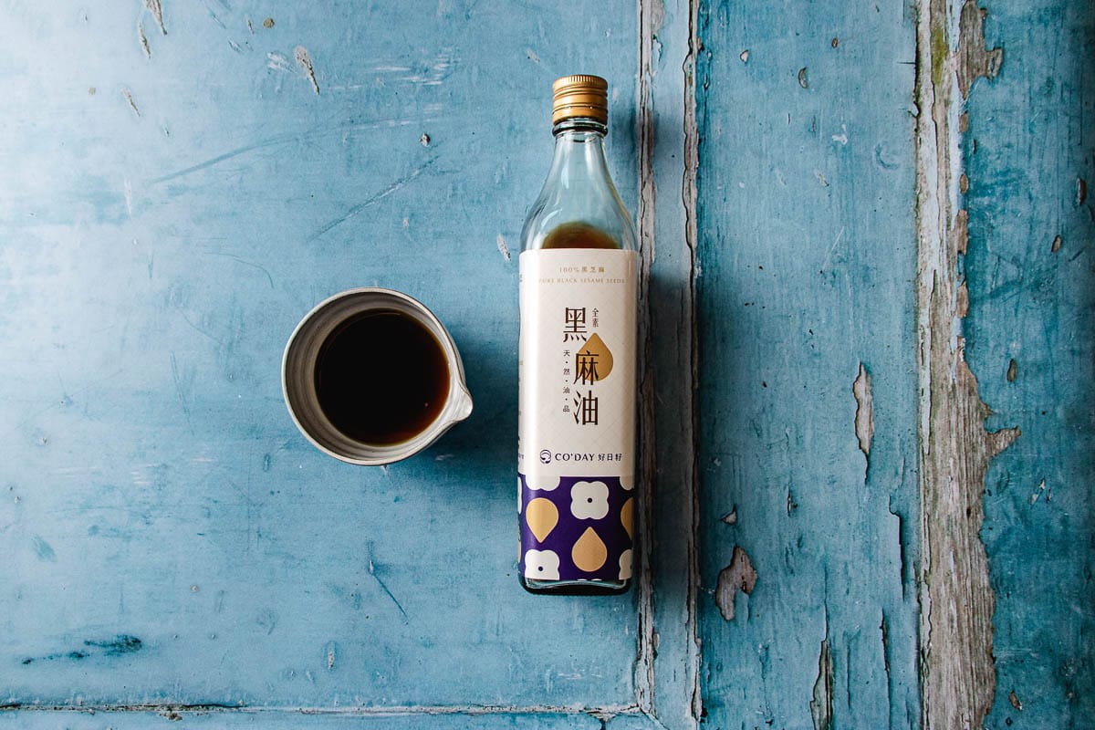 A bottle of Taiwanese black sesame oil with a blue and gold label sits next to a small ceramic cup filled with dark liquid—perfect for enhancing sesame oil chicken soup—on a weathered blue wooden surface.
