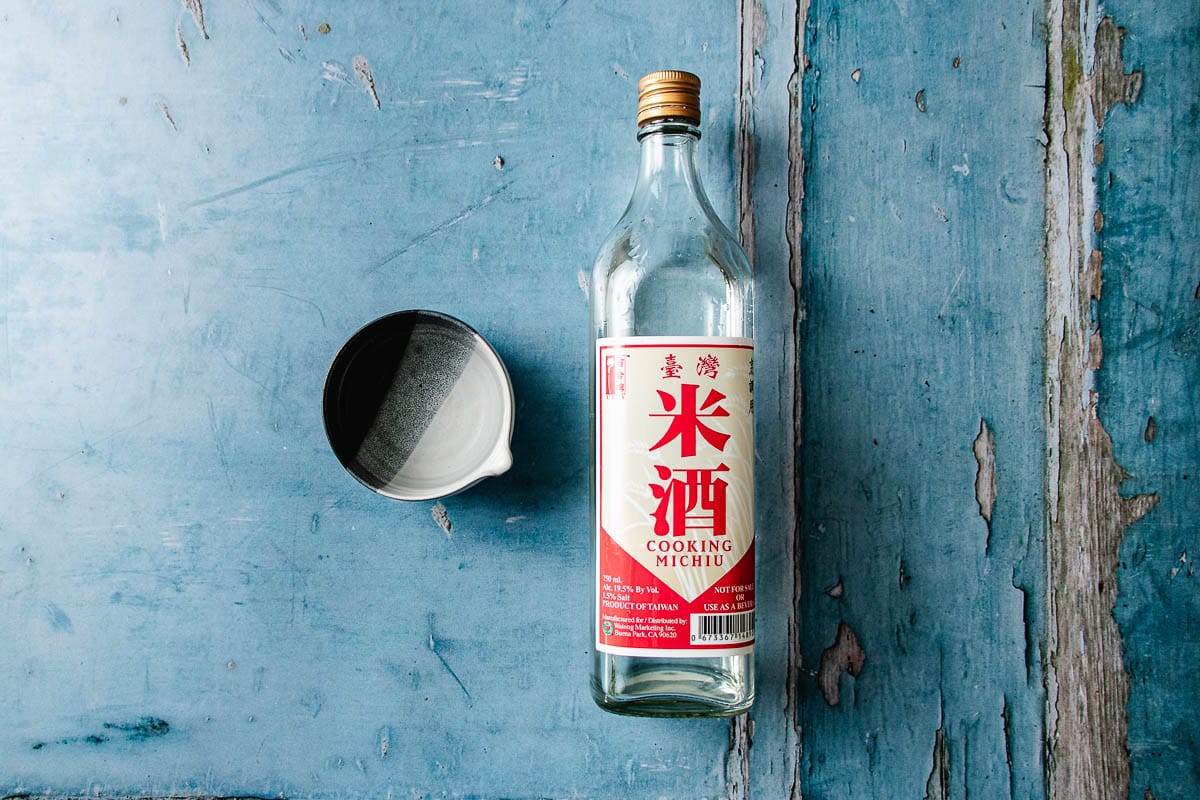 A clear bottle of Michiu cooking wine with a red and white label sits next to a small empty white bowl on a distressed blue wooden surface, perfect for preparing flavorful dishes like sesame oil chicken soup.