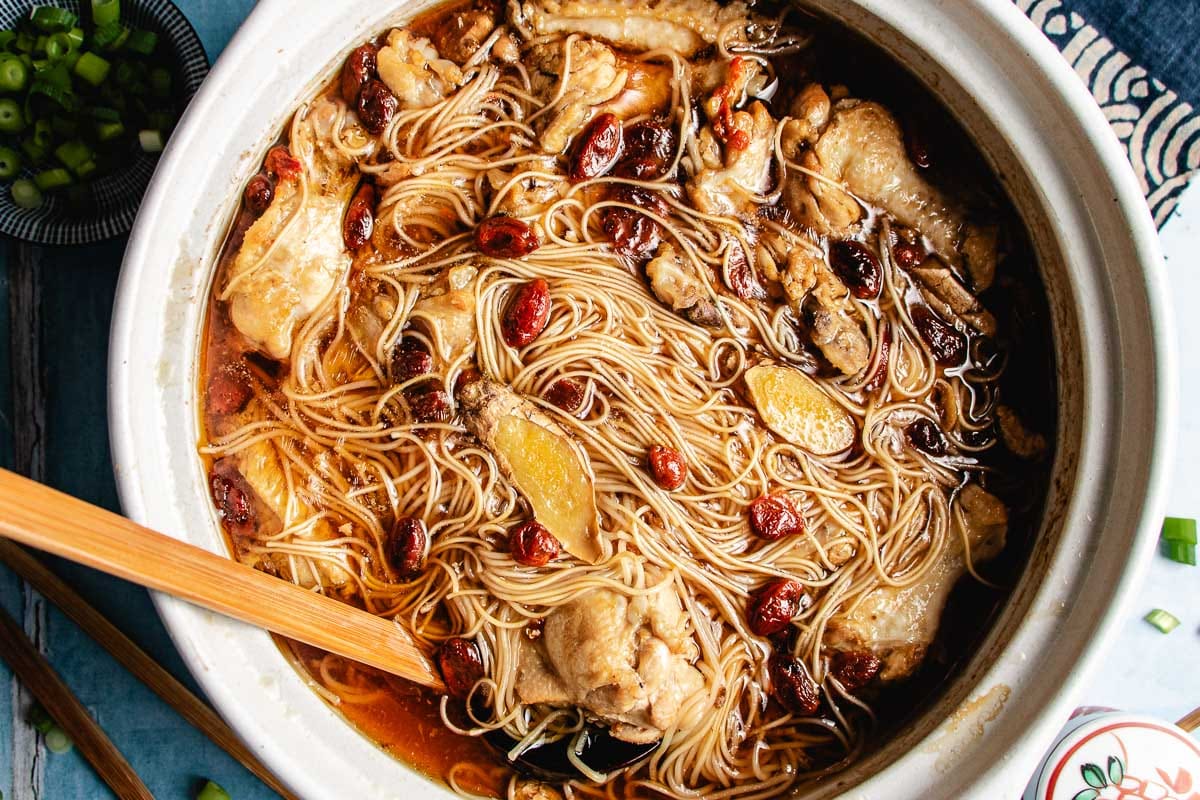 A bowl of sesame oil chicken soup with cut-up chicken pieces, sliced ginger, dried goji berries, and a rich brown broth, served in a large ceramic pot with a wooden spoon.