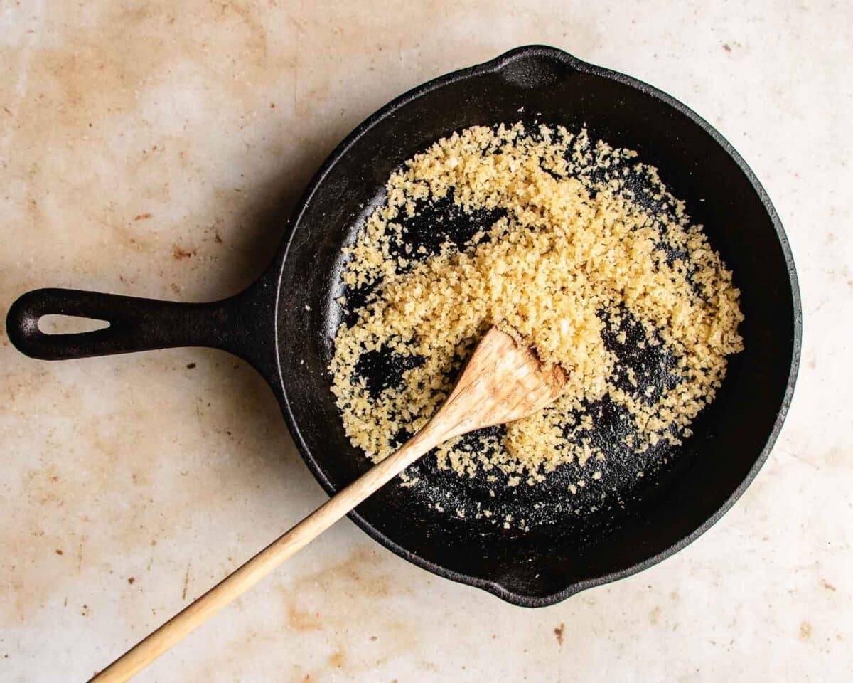 A cast iron skillet containing toasted breadcrumbs for roasted napa cabbage sits on a light countertop. A wooden spatula rests in the skillet, partially mixing the breadcrumbs.