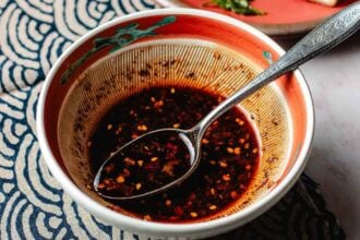 A bowl of spicy wonton/dumpling dipping sauce with chili flakes and seeds, perfect for Smashed Dumpling Tacos, featuring a metal spoon resting inside. The bowl has a decorative interior and sits on a patterned blue and white cloth.