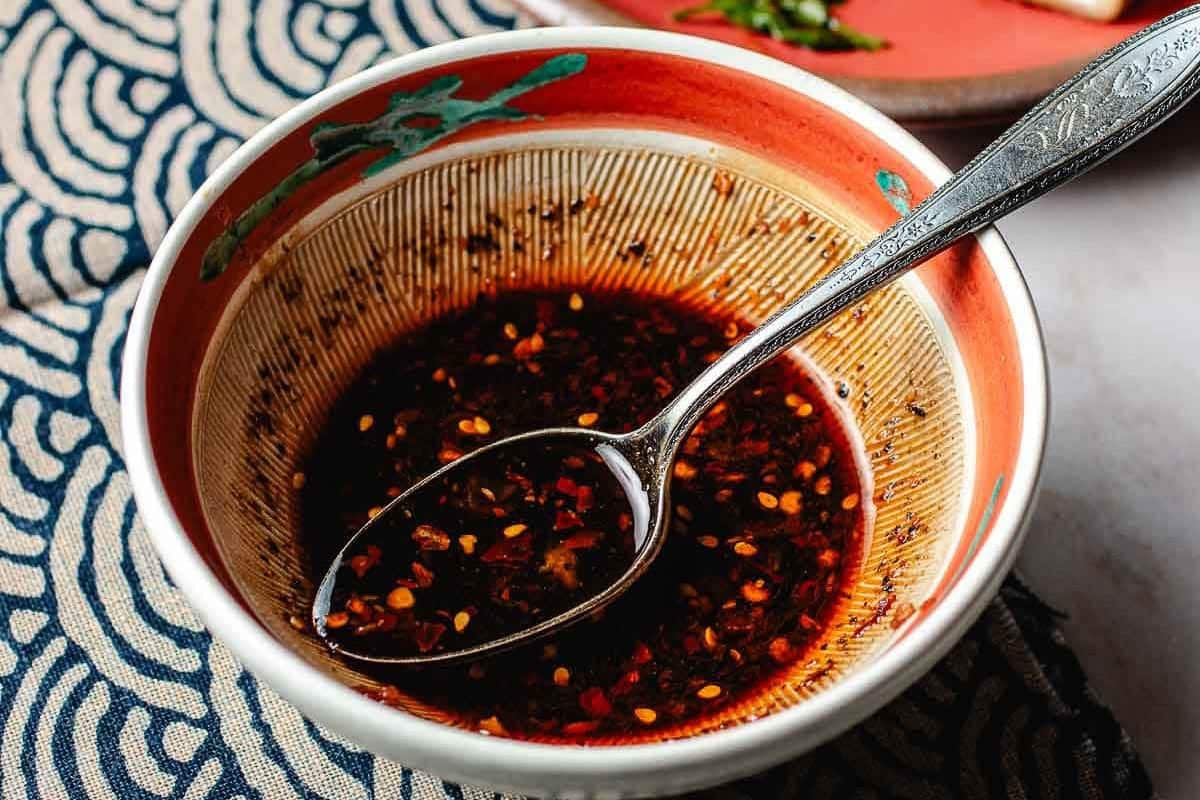 A bowl of spicy wonton/dumpling dipping sauce with chili flakes and seeds, perfect for Smashed Dumpling Tacos, featuring a metal spoon resting inside. The bowl has a decorative interior and sits on a patterned blue and white cloth.