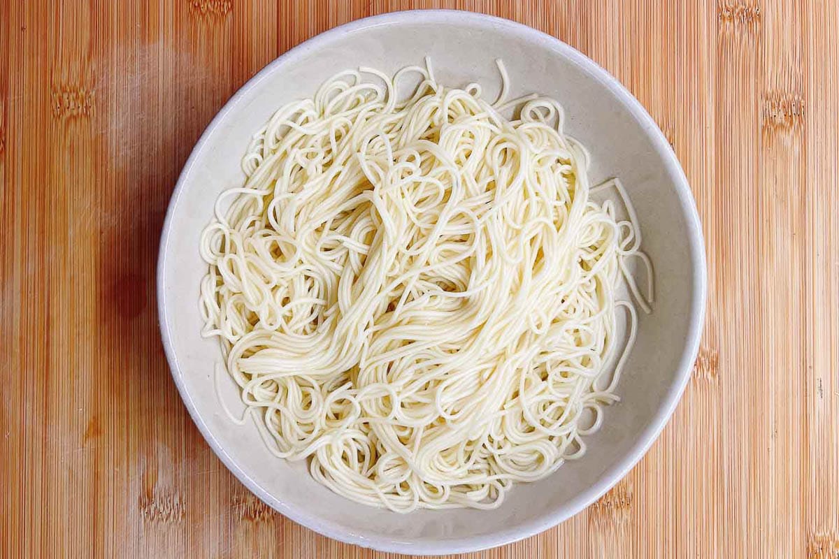 A bowl of plain, cooked white noodles sits on a wooden surface, ready to be transformed into a comforting egg drop noodle soup.