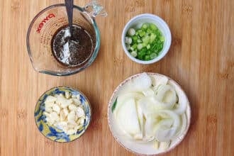Four bowls on a wooden surface featuring ingredients for Bulgogi Fries: sliced green onions, sliced garlic, sliced white onions, and a measuring cup with rich bulgogi sauce and a spoon.