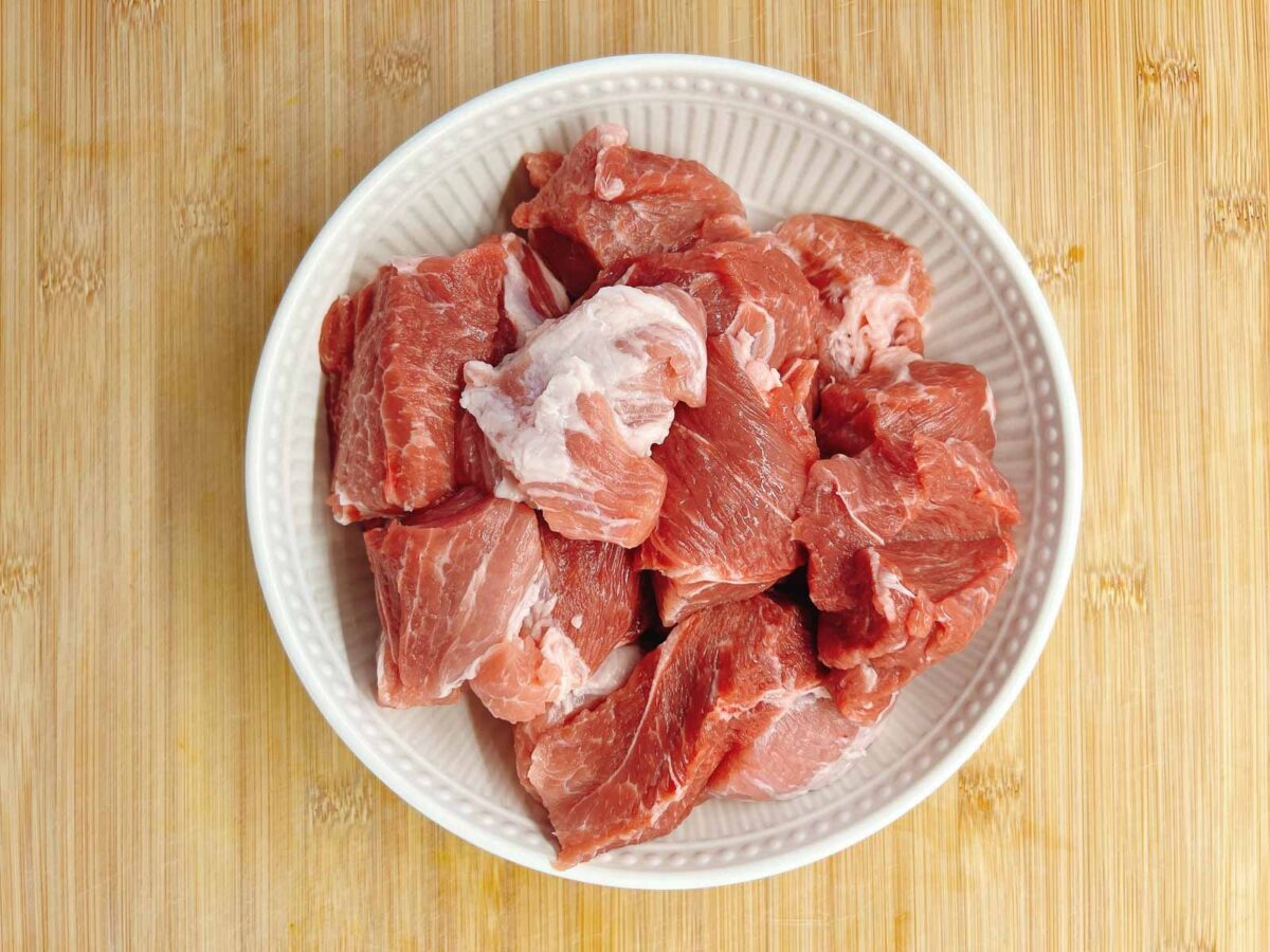 A white bowl filled with raw, cut pieces of red meat—perfect for preparing Mongolian Pork Slow Cooker recipes—rests on a light wooden surface.