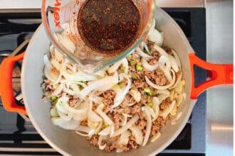 A pot with browned ground meat, sliced onions, and green onions is on a stovetop. A hand is pouring a dark bulgogi sauce from a glass measuring cup into the pan—perfect for making savory Bulgogi Fries.