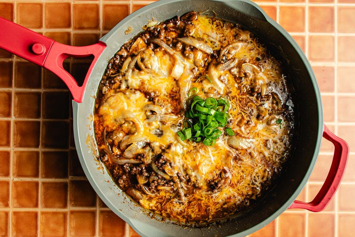 A skillet filled with cheesy bulgogi beef and onion mixture, inspired by Bulgogi Fries, is topped with melted cheese and sliced green onions, sitting on a red tiled countertop.
