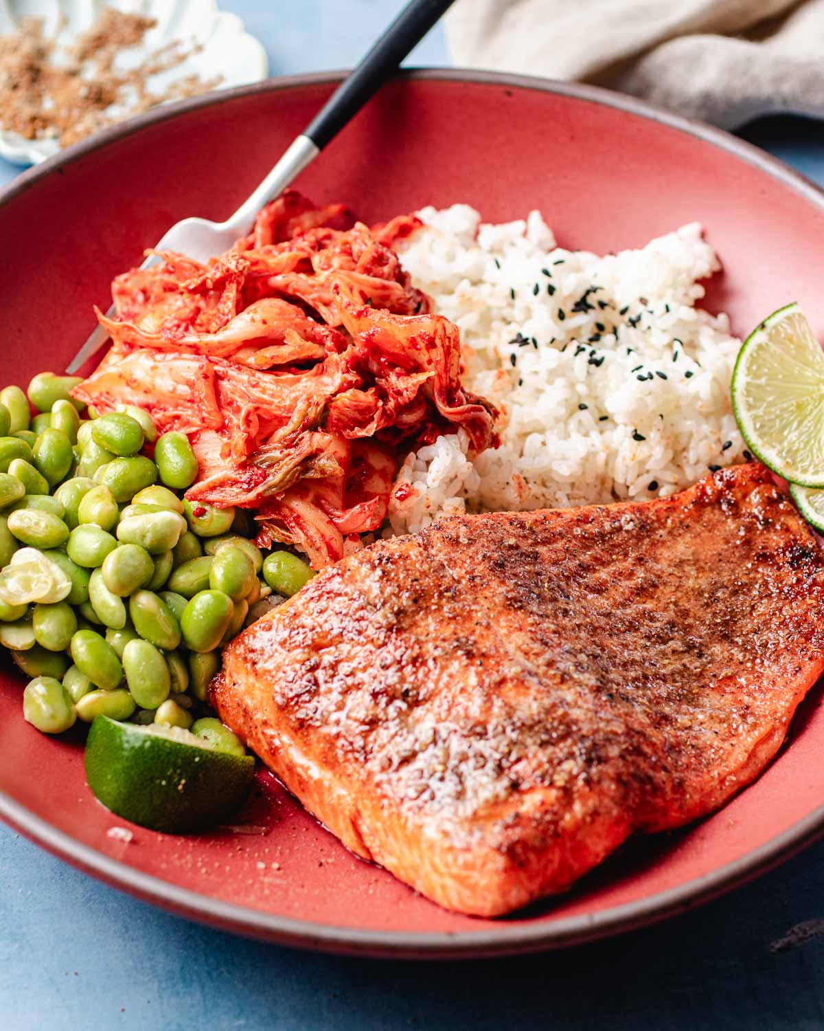 A bowl with air fried trout fillet, white rice topped with black sesame seeds, kimchi, edamame, and lime wedges. A fork rests near the kimchi on the red plate.