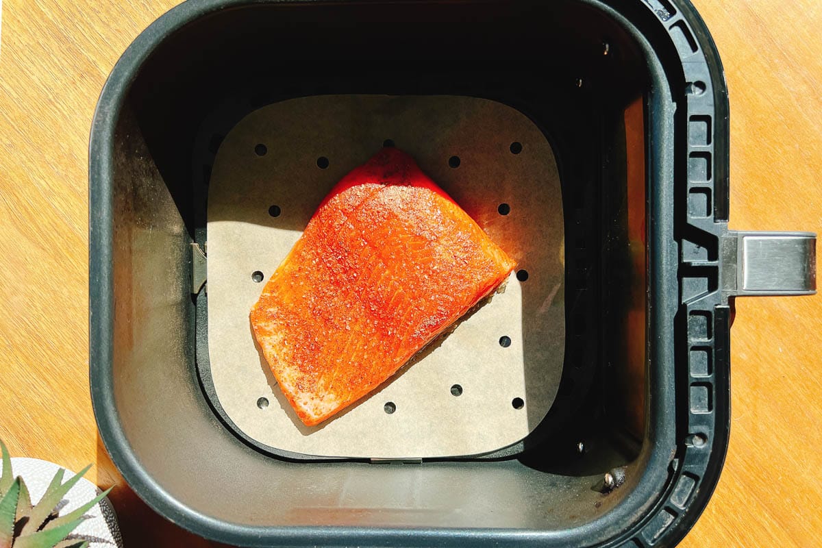 A piece of seasoned trout fillet sits on parchment paper inside an air fryer basket, ready to be cooked—just like preparing air fryer trout. Sunlight shines on the air fryer from the left side.