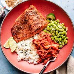 A plate with a serving of air fryer trout, steamed white rice with black sesame seeds, edamame, kimchi, and lime wedges, arranged on a reddish-brown dish with a fork resting by the kimchi.