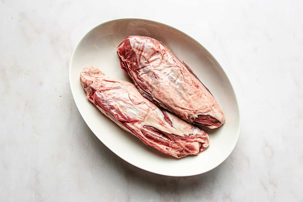 Two raw pieces of beef shank, boneless, placed side by side on an oval white plate against a light-colored surface.