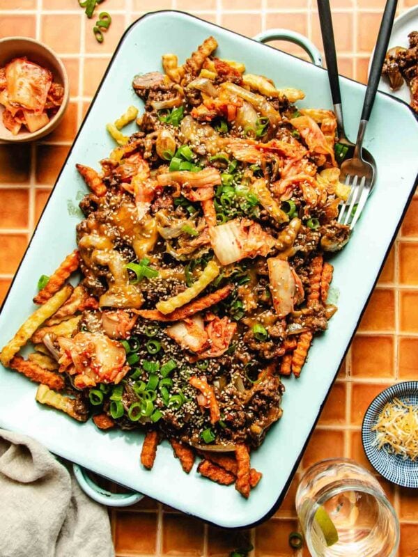A tray of Bulgogi Fries—crinkle-cut fries topped with ground beef, kimchi, green onions, and sesame seeds—is served with a fork and spoon. Small bowls of kimchi and grated cheese sit nearby on a tiled surface.