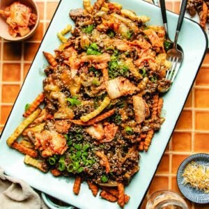 A platter of Bulgogi Fries featuring crinkle-cut fries topped with kimchi, green onions, sesame seeds, shredded cheese, and seasoned beef on a light blue tray, served with a fork and knife on the side.