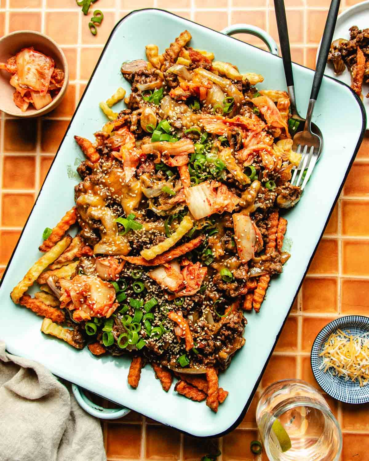 A tray of Bulgogi Fries—crinkle-cut fries topped with ground beef, kimchi, green onions, and sesame seeds—is served with a fork and spoon. Small bowls of kimchi and grated cheese sit nearby on a tiled surface.