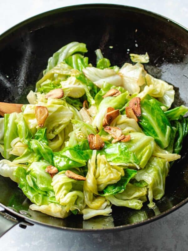 Taiwanese cabbage with slices of crispy garlic chips in a black wok, with a wooden spatula partially visible on the left side. Reminiscent of Din Tai Fung recipe, the cabbage is bright green and slightly wilted.