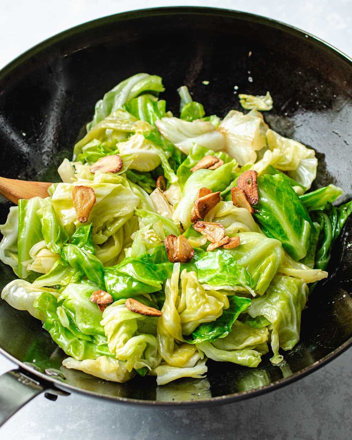 Taiwanese cabbage with slices of crispy garlic chips in a black wok, with a wooden spatula partially visible on the left side. Reminiscent of Din Tai Fung recipe, the cabbage is bright green and slightly wilted.