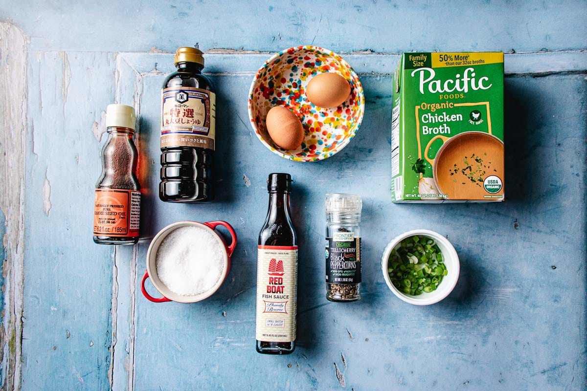 A flat lay for egg drop noodle soup ingredients on a blue surface: soy sauce, eggs in a colorful bowl, chicken broth, green onions in a small bowl, ground black pepper, fish sauce, in a red cup, and oyster sauce.