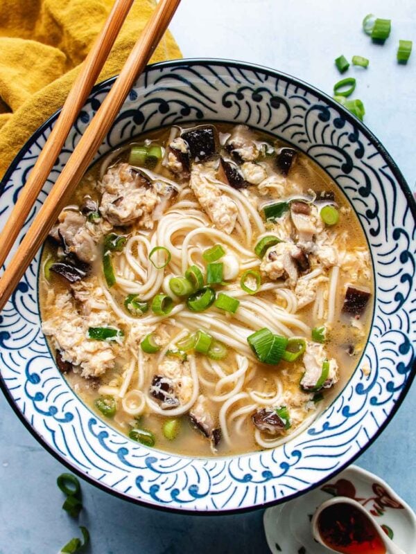 A bowl of egg drop noodle soup with shiitake, green onions, and delicate strands of egg in a patterned dish, garnished with chopped scallions and accompanied by wooden chopsticks.