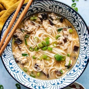 A bowl of egg drop noodle soup with chopped green onions, mushrooms, and bits of egg in a patterned bowl, with chopsticks resting on the rim and a yellow cloth nearby.