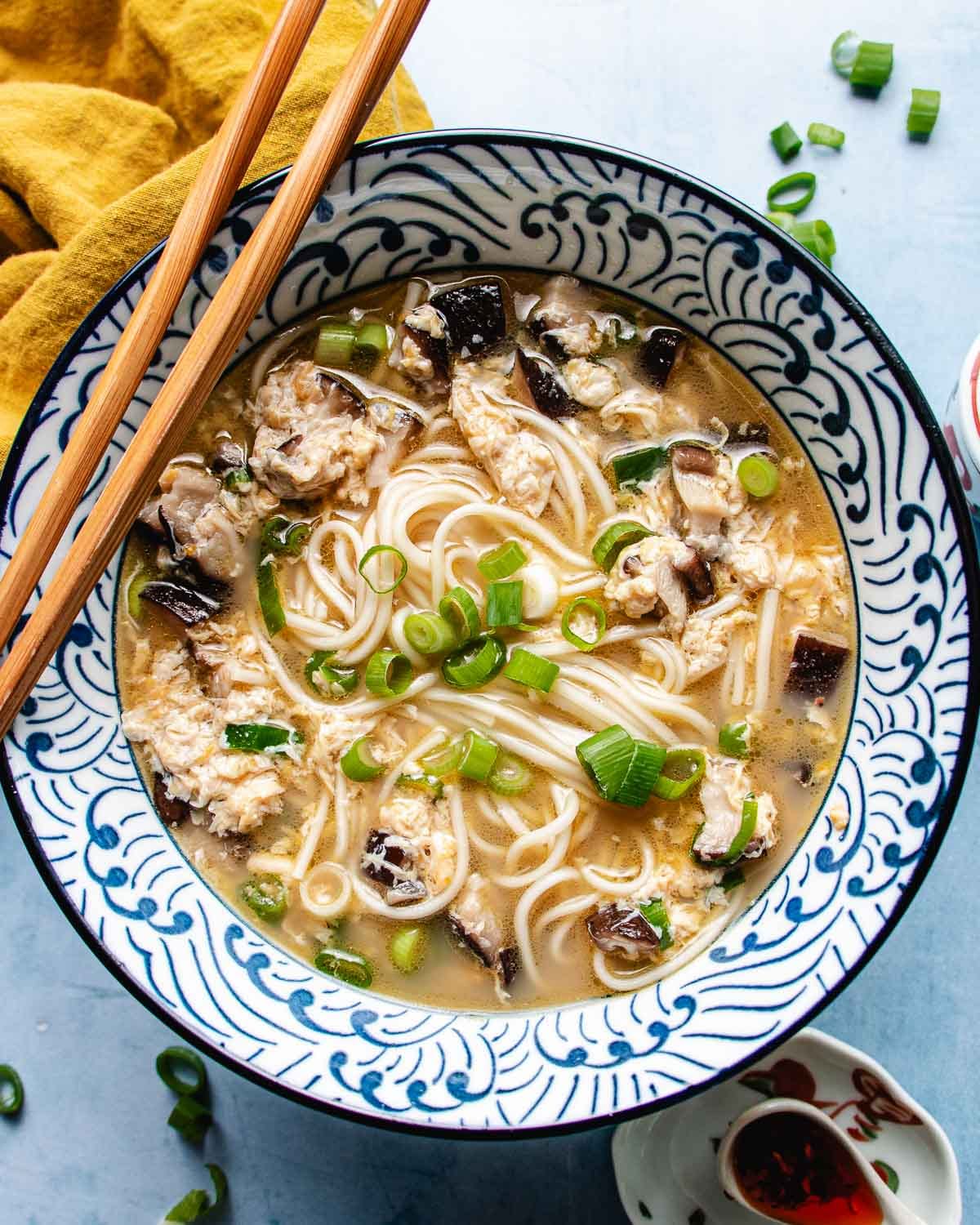 A bowl of egg drop noodle soup with shiitake, green onions, and delicate strands of egg in a patterned dish, garnished with chopped scallions and accompanied by wooden chopsticks.