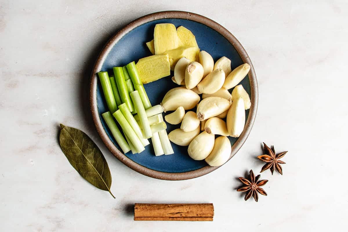 A blue plate holds sliced ginger, whole garlic cloves, and chopped green onions—essential aromatics for making Taiwanese Braised Beef Shank. Nearby are a bay leaf, two star anise pods, and a cinnamon stick on a light surface.