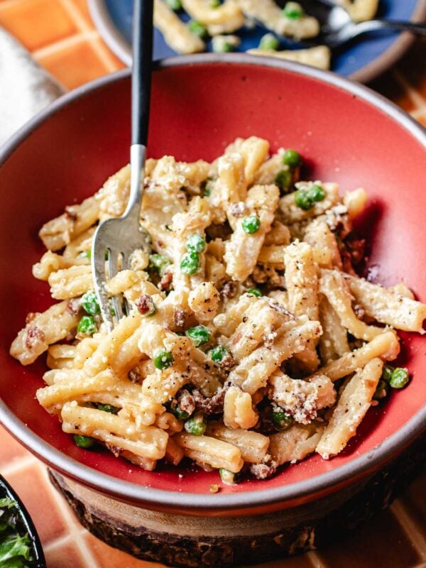 A bowl of creamy miso butter pasta with green peas and bits of pancetta/bacon, topped with grated cheese and black pepper, with a fork resting in the bowl.