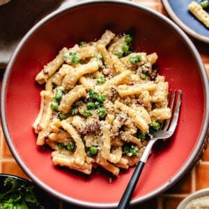 A bowl of creamy miso butter pasta mixed with green peas and sprinkled with crispy pancetta bits, black pepper and grated cheese, served with a fork on a red plate.